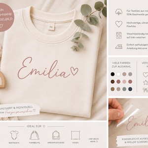 Könnte beinhalten: Cremefarbenes Sweatshirt mit dem Namen "Emilia" in Schreibschrift und einem Herzsymbol. Das Sweatshirt hat einen Rundhalsausschnitt und ein Etikett mit der Nummer "98". Das Bild enthält auch deutschen Text.