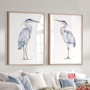 May include: Two framed watercolor prints featuring a blue heron. The birds are depicted in profile, with long legs and beaks, set against a white background. The frames are a light wood color. The text "Frames are NOT INCLUDED" is at the bottom.