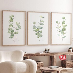 May include: Three framed watercolor prints featuring eucalyptus branches with green and blue-green leaves. The prints are in light brown wooden frames and are displayed above a wooden console table. The room has a neutral color palette.