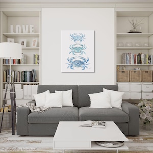 May include: A vertical canvas print featuring three blue crab illustrations. The crabs are in varying shades of blue and are arranged one above the other. The artwork is displayed on a white wall above a gray sofa in a living room setting.