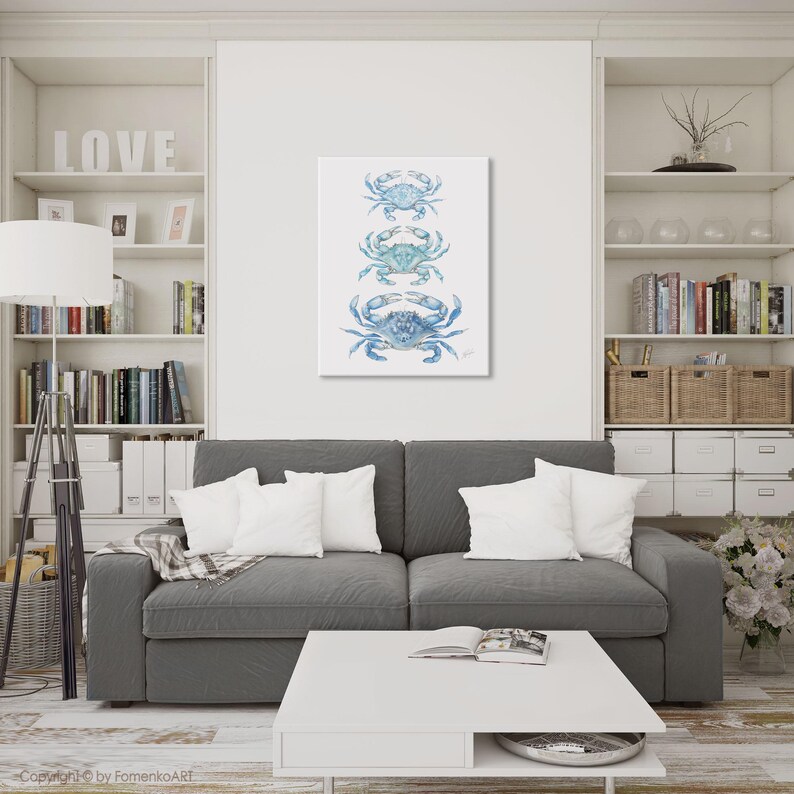 May include: A vertical canvas print featuring three blue crab illustrations. The crabs are in varying shades of blue and are arranged one above the other. The artwork is displayed on a white wall above a gray sofa in a living room setting.