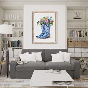 May include: A framed watercolor print featuring blue cowboy boots filled with colorful flowers. The boots have a detailed blue and white floral pattern. The artwork is displayed in a living room setting with a gray sofa and white shelves.