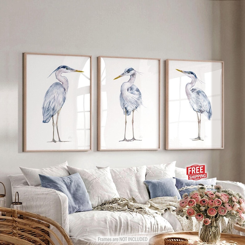 May include: A set of three watercolor paintings featuring blue heron birds on a white background. The paintings are framed in a light wood frame.