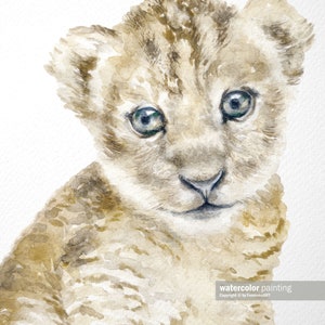 May include: A watercolor painting of a light brown lion cub with blue eyes. The cub is looking directly at the viewer. The painting is on a white background.