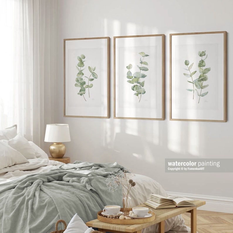 Puede incluir: Tres acuarelas enmarcadas de ramas de eucalipto con hojas verdes sobre un fondo blanco. Las pinturas est&aacute;n colgadas en una pared blanca en un dormitorio.