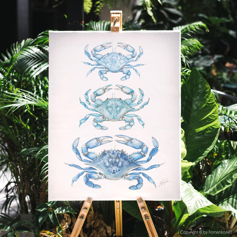 May include: A watercolor painting of three blue crabs arranged vertically on a white canvas. The crabs are in varying shades of blue, with detailed depictions of their shells and claws. The artwork is displayed on a wooden easel, with lush green foliage in the background.