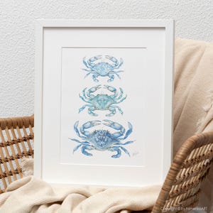 May include: A framed art print featuring three blue crab illustrations in varying shades of blue. The crabs are arranged vertically on a white background, set within a white frame. The artwork has a watercolor style and evokes a coastal theme.
