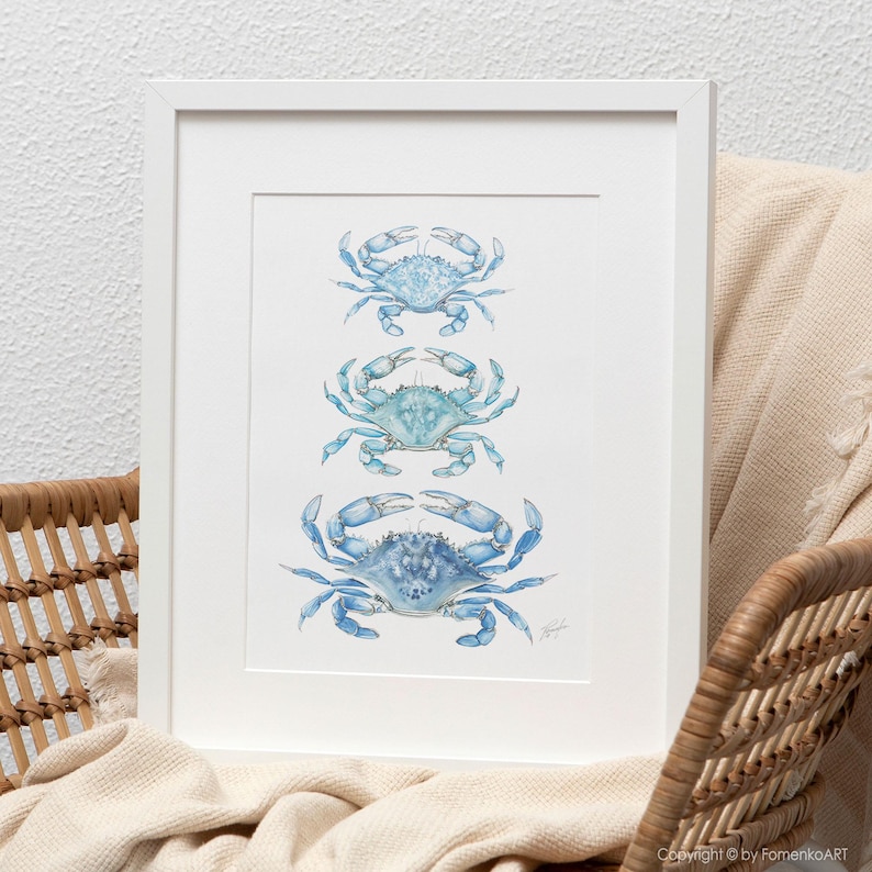 May include: A framed art print featuring three blue crab illustrations in varying shades of blue. The crabs are arranged vertically on a white background, set within a white frame. The artwork has a watercolor style and evokes a coastal theme.