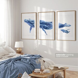 May include: Three framed watercolor paintings of blue whales. The paintings are in a modern style and feature a mother whale and her calf. The paintings are hung on a white wall in a bedroom setting.