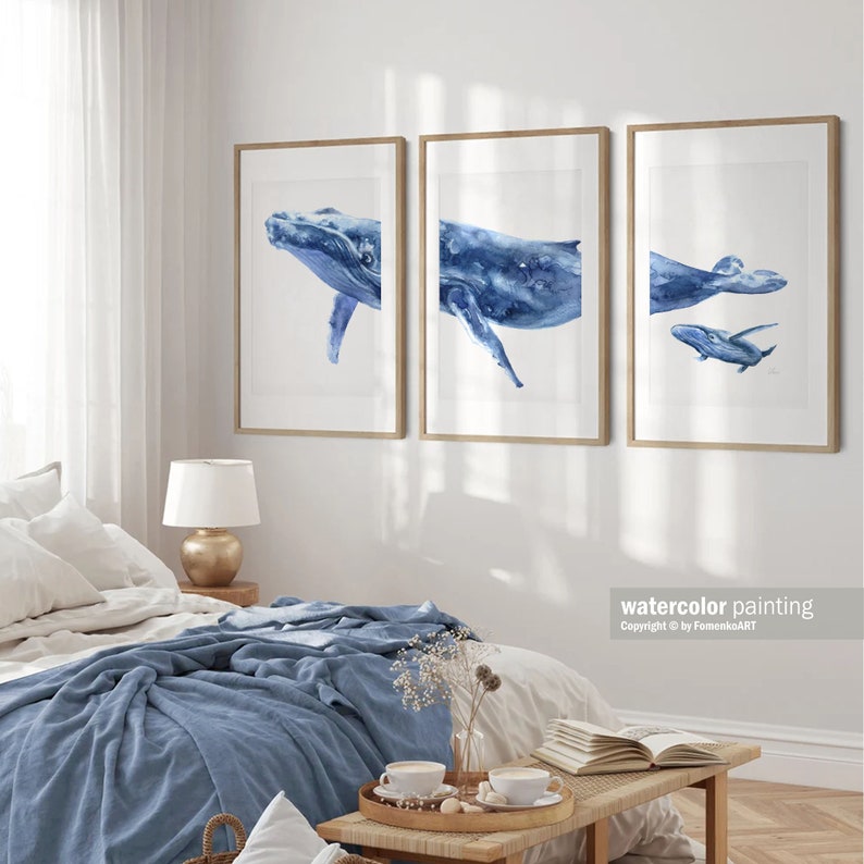 May include: Three framed watercolor paintings of blue whales. The paintings are in a modern style and feature a mother whale and her calf. The paintings are hung on a white wall in a bedroom setting.