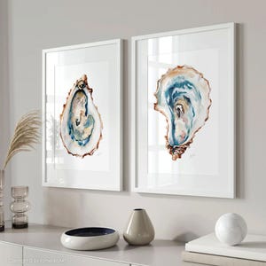 Puede incluir: Dos impresiones enmarcadas en acuarela de conchas de ostra. La obra de arte presenta tonos de azul, marr&oacute;n y blanco, con detalles intrincados. Las impresiones se exhiben en marcos blancos, agregando un toque de elegancia al arte de tem&aacute;tica costera.