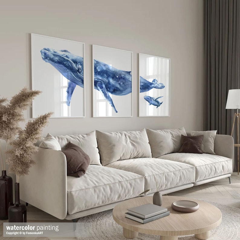 May include: Three framed watercolor paintings of blue whales. The whales are depicted in various poses, with one showing the whale's underside. The paintings are displayed above a beige sofa in a living room setting. The text "watercolor painting" is visible.