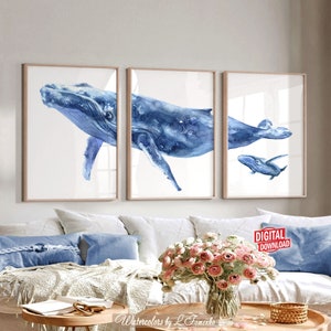 May include: Three framed watercolor prints of whales in shades of blue. The artwork features a large whale, a medium-sized whale, and a baby whale. The prints are displayed above a white couch with blue pillows and a coffee table with flowers.