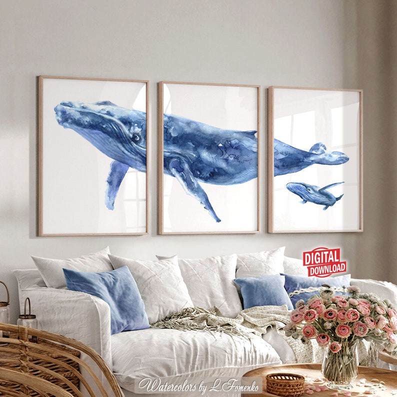 May include: Three framed watercolor prints of whales in shades of blue. The artwork features a large whale, a medium-sized whale, and a baby whale. The prints are displayed above a white sofa with blue pillows and a wooden coffee table.