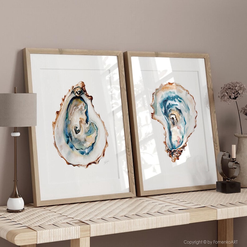 Puede incluir: Dos impresiones enmarcadas en acuarela de conchas de ostra. La obra de arte presenta tonos de azul, marr&oacute;n y blanco, con una representaci&oacute;n detallada del interior de la concha. Los marcos son de madera clara y el arte se exhibe sobre una superficie clara.