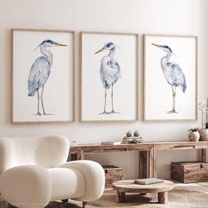 May include: Three watercolor paintings of grey herons with yellow beaks, each in a separate wooden frame, hanging on a white wall.