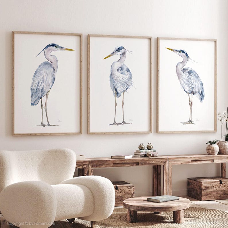 May include: Three watercolor paintings of grey herons with yellow beaks, each in a separate wooden frame, hanging on a white wall.