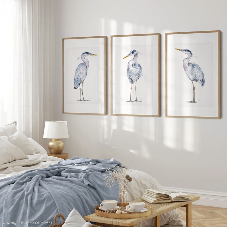 May include: Three framed watercolor paintings of blue herons, each with a different pose, on a white wall in a bedroom setting.