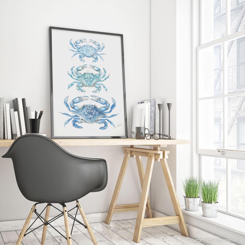 May include: A framed art print featuring three blue crab illustrations in varying shades of blue. The crabs are arranged vertically, with a black frame. The artwork is displayed on a wooden desk with a black chair.