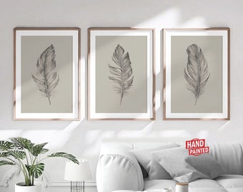 May include: Three framed prints of hand-drawn feather illustrations on a beige background. The feathers are drawn in shades of gray and are arranged vertically in the center of each print. The prints are displayed on a white wall above a white couch.