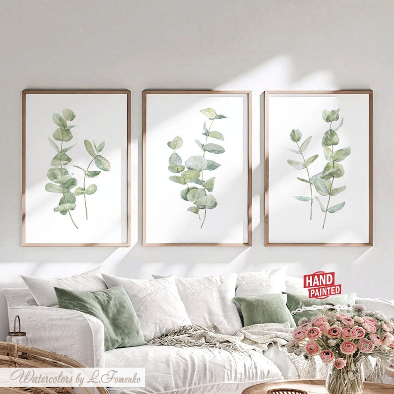 Pu&ograve; includere: Set di tre stampe ad acquerello incorniciate con foglie di eucalipto verdi su sfondo bianco. Le stampe sono in semplici cornici di legno e sono appese a una parete bianca sopra un divano bianco.