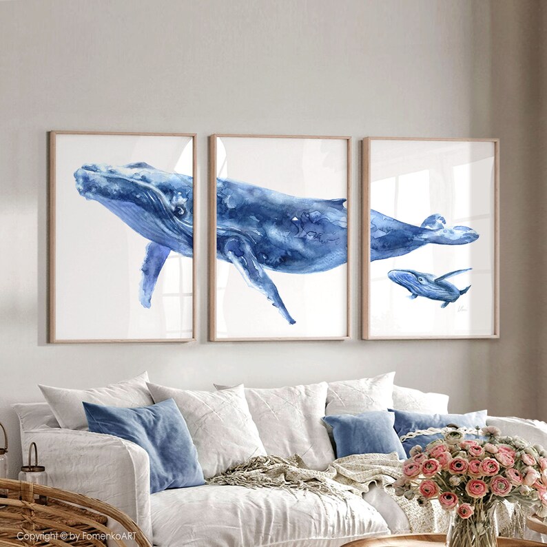 May include: A set of three watercolor paintings featuring blue whales. The paintings are framed and displayed on a white wall above a white couch with blue pillows.