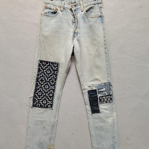 Può includere: Jeans di denim azzurro con toppe. I jeans hanno una chiusura con bottoni e sono appesi a una gruccia di legno. Le toppe sono cucite sulle ginocchia, con disegni geometrici e fantasia. I jeans mostrano segni di usura.