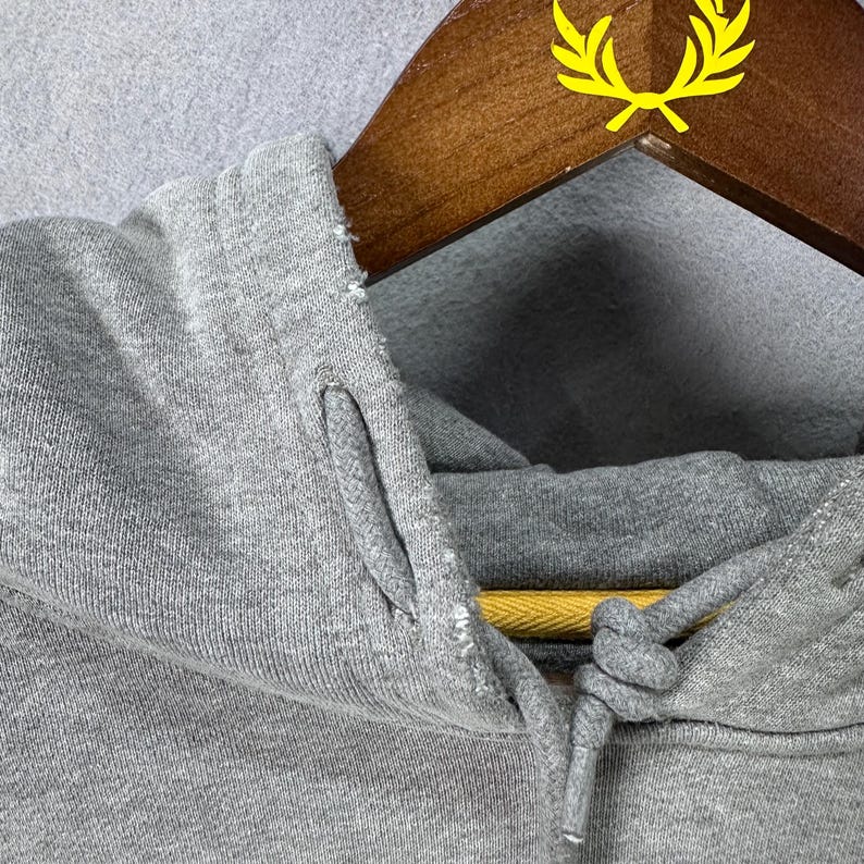 May include: Close-up of a gray hoodie with a drawstring and a hood. The hoodie is hanging on a wooden hanger with a yellow logo. The fabric appears to be a soft, knit material.