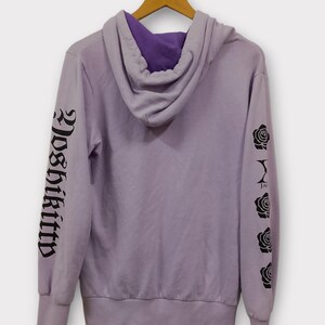 Pu&ograve; includere: Una felpa con cappuccio color lavanda con cappuccio foderato di viola. La manica sinistra presenta la scritta "Yoshikitty" in caratteri gotici neri, mentre la manica destra mostra grafiche di rose nere. La felpa &egrave; appesa a una gruccia di legno.