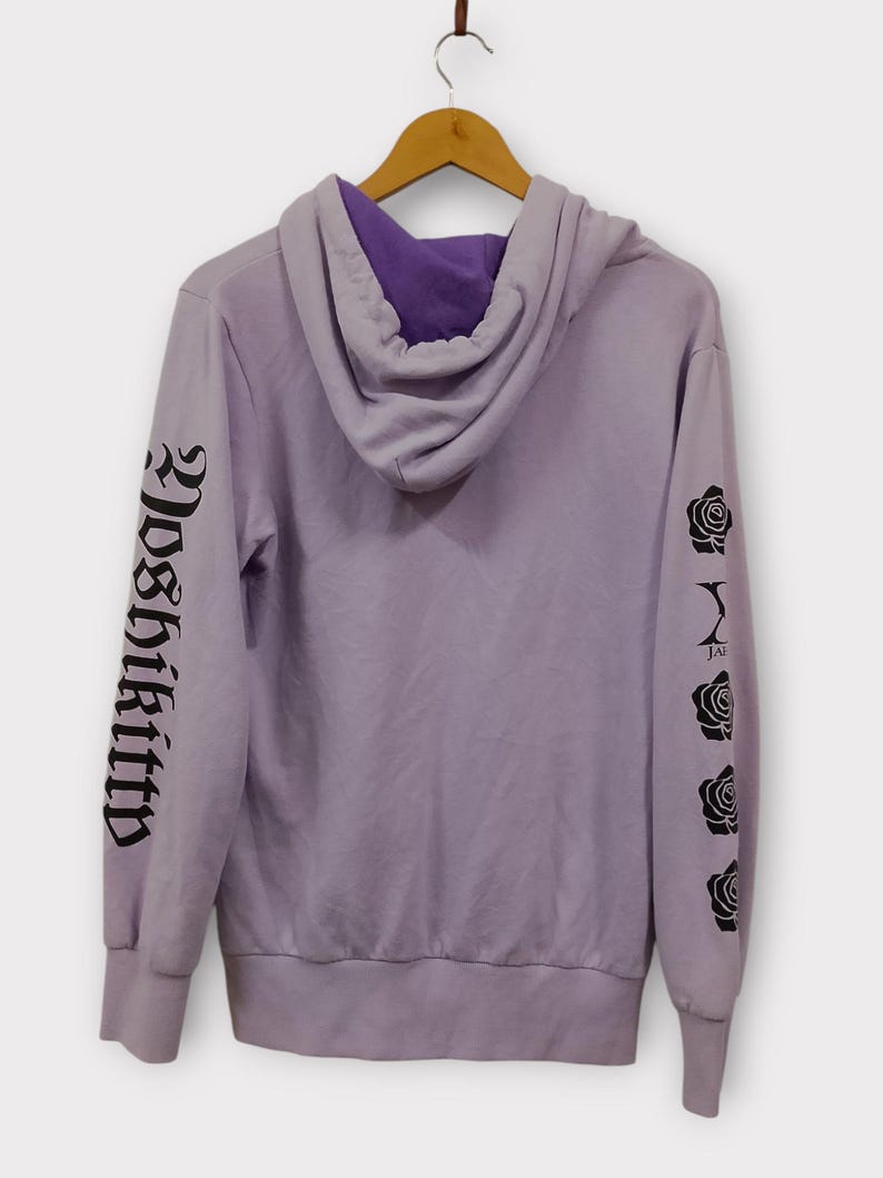 Pu&ograve; includere: Una felpa con cappuccio color lavanda con cappuccio foderato di viola. La manica sinistra presenta la scritta "Yoshikitty" in caratteri gotici neri, mentre la manica destra mostra grafiche di rose nere. La felpa &egrave; appesa a una gruccia di legno.