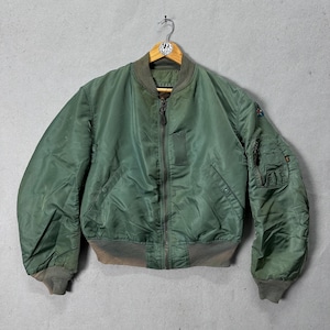 May include: Green bomber jacket with a zip-up front, ribbed cuffs, and a collar. The jacket has a pocket on the left sleeve and is hanging on a wooden hanger. The jacket is a solid green colour.
