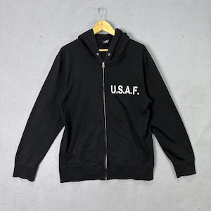 Könnte beinhalten: Schwarzer Kapuzenpullover mit Reißverschluss, Kapuze und Vordertaschen. Der weiße Text "U.S.A.F." ist auf der Vorderseite aufgedruckt. Der Hoodie hängt an einem Holzbügel vor grauem Hintergrund.