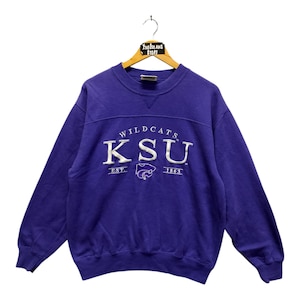 May include: A purple sweatshirt with the text "Wildcats KSU" in white, with a stylized wildcat logo. The sweatshirt has a crew neck and long sleeves. The text "EST. 1863" is below the main text. The sweatshirt is hanging on a wooden hanger.