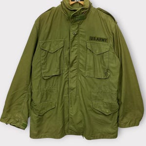 May include: Olive green military jacket with multiple front pockets and the text "U.S. ARMY" on the chest. The jacket has a high collar and is hanging on a hanger. A vintage piece.
