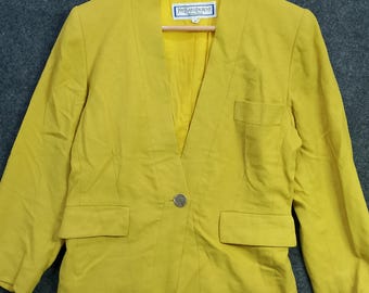 May include: A mustard yellow blazer with a notched collar, two front pockets, and a single button closure. The blazer is hanging on a wooden hanger. The label reads "Yves Saint Laurent".