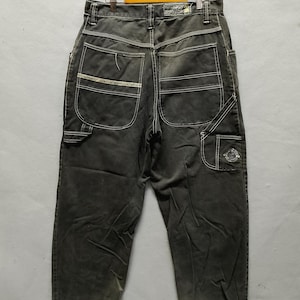 May include: Dark gray denim jeans with white stitching, visible pockets, and a distressed hem. The jeans are hanging on a wooden hanger. The back pockets have a decorative design. The jeans have a relaxed fit.