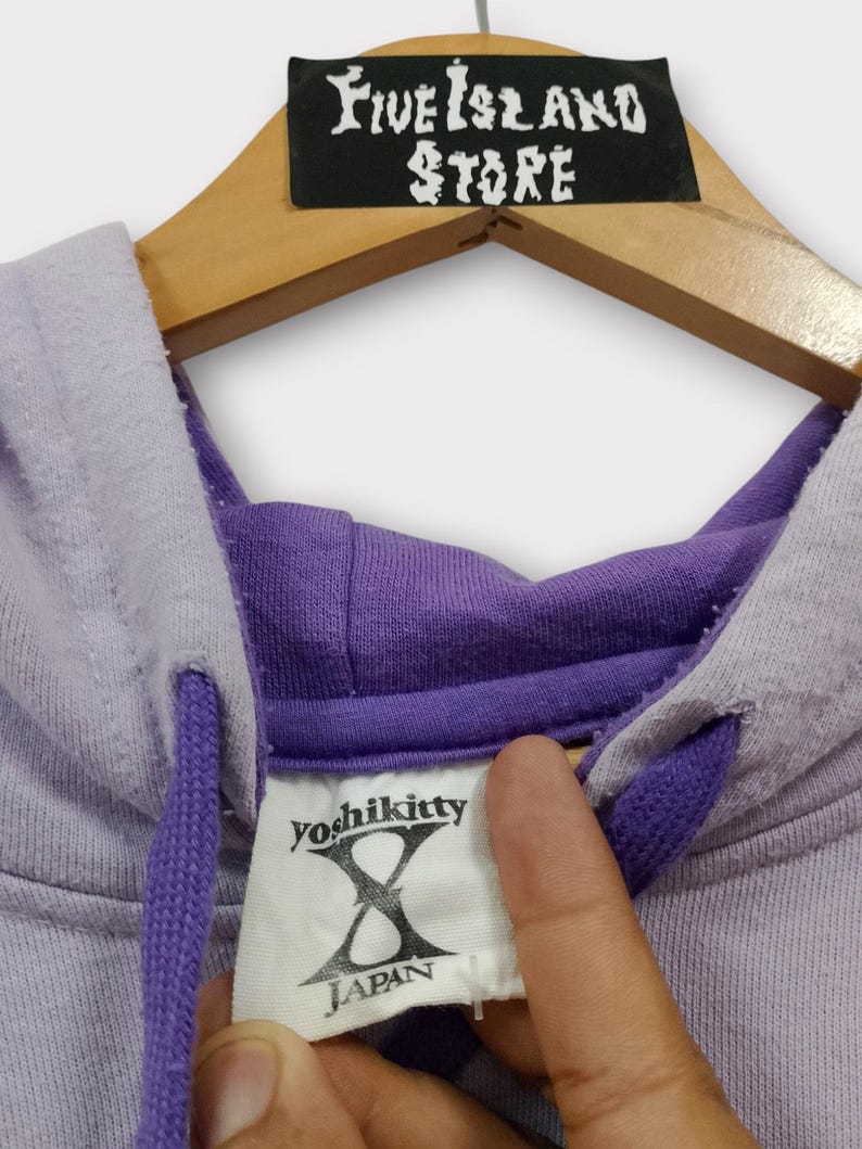 Pu&ograve; includere: Una felpa con cappuccio lilla con coulisse viola e cappuccio interno viola. L'etichetta recita "yoshikitty X JAPAN". La felpa con cappuccio &egrave; appesa a una gruccia di legno con un'etichetta nera che dice "Five Island Store".