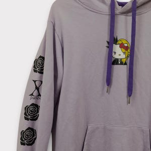 Pu&ograve; includere: Una felpa con cappuccio lavanda con una tasca frontale e un cappuccio con coulisse viola. La manica sinistra presenta grafiche di rose nere e la scritta "JAPAN". Un personaggio dei cartoni animati con un fiocco &egrave; sul davanti.