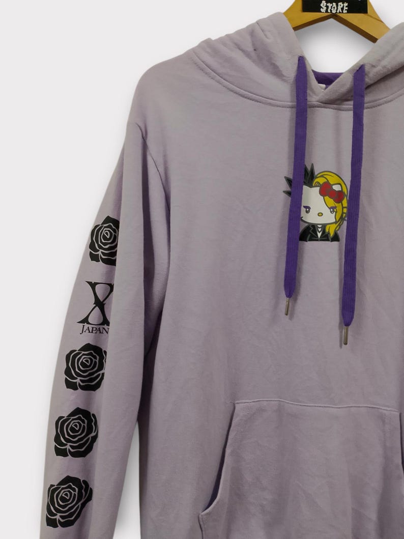 Pu&ograve; includere: Una felpa con cappuccio lavanda con una tasca frontale e un cappuccio con coulisse viola. La manica sinistra presenta grafiche di rose nere e la scritta "JAPAN". Un personaggio dei cartoni animati con un fiocco &egrave; sul davanti.