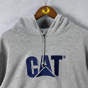 May include: A gray hooded sweatshirt with the CAT logo in navy blue. The sweatshirt has a drawstring and is hanging on a wooden hanger.