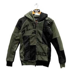 Peut inclure: Un sweat à capuche zippé en patchwork vert et noir. Il présente une capuche, une fermeture éclair intégrale et une poche avant. Le design comprend du texte et diverses textures de tissu. Le vêtement est suspendu à un cintre en bois.
