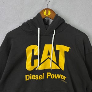 May include: Black hooded sweatshirt featuring the CAT logo in yellow. The words "Diesel Power" are printed below the logo in yellow. The sweatshirt has a drawstring and a kangaroo pocket. The sweatshirt is hanging on a wooden hanger.