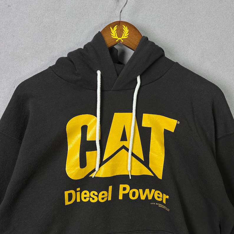 May include: Black hooded sweatshirt featuring the CAT logo in yellow. The words "Diesel Power" are printed below the logo in yellow. The sweatshirt has a drawstring and a kangaroo pocket. The sweatshirt is hanging on a wooden hanger.