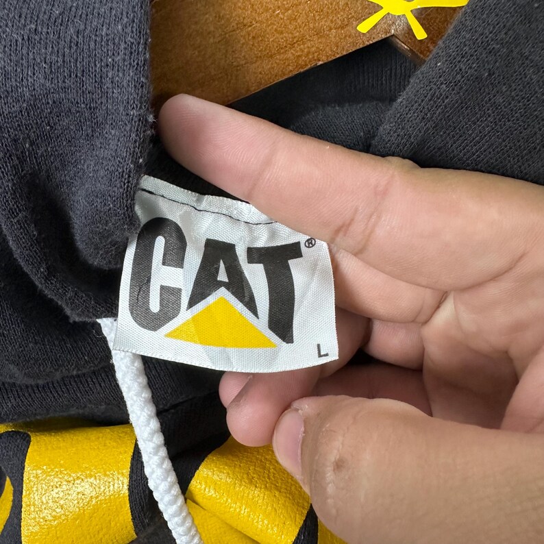 May include: Close-up of a black hoodie tag with the word "CAT" in bold, black letters. The "A" in "CAT" is a yellow triangle. The tag is white with a black border. The hoodie has a white drawstring.