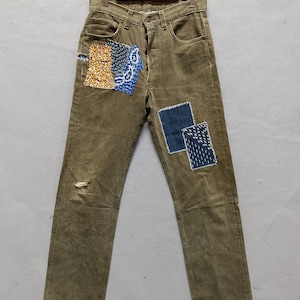 Può includere: Jeans in denim verde oliva con toppe decorative. Le toppe sono nelle tonalità dell'arancione, del blu e del blu navy. I jeans hanno un aspetto invecchiato con un piccolo strappo sul ginocchio sinistro. I jeans sono appesi a una gruccia di legno.