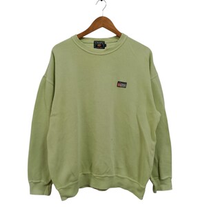 May include: A pale green Chaps sweatshirt with a crew neck and long sleeves. The sweatshirt features a small logo patch on the chest and a larger logo on the back of the neck. The garment is suspended from a wooden hanger.