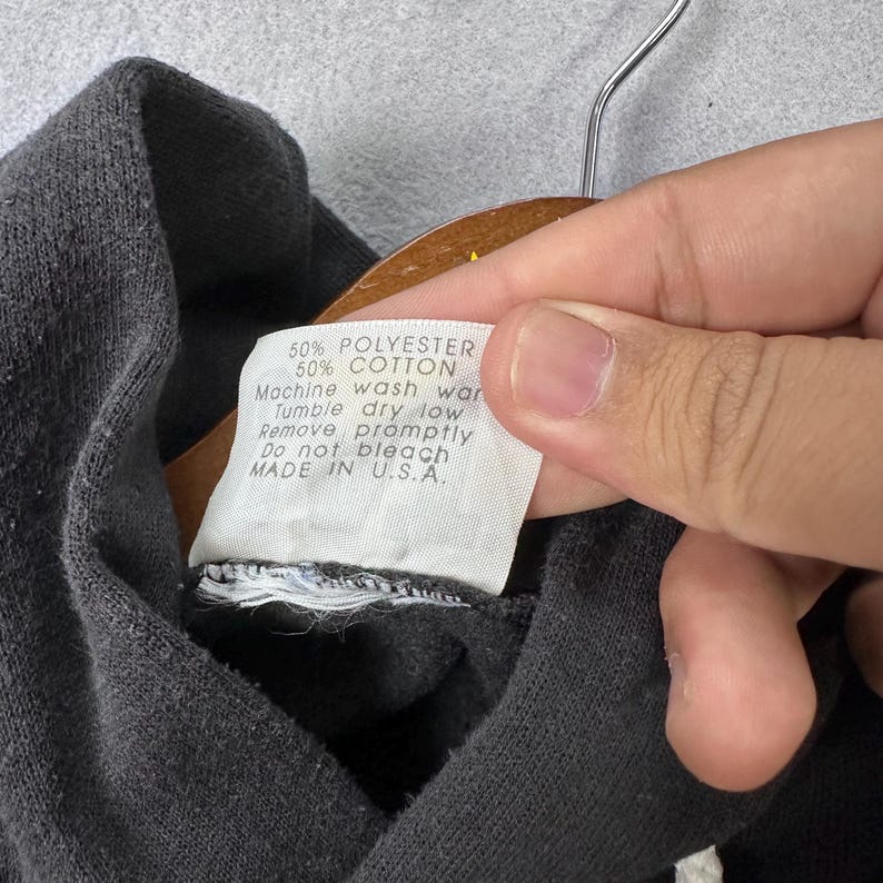 May include: A close-up of a dark grey sweatshirt collar with a label. The label states "50% POLYESTER 50% COTTON Machine wash war Tumble dry low Remove promptly Do not bleach MADE IN U.S.A."