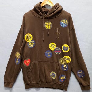 Puede incluir: Sudadera con capucha marrón con múltiples parches circulares. Estos parches muestran texto y gráficos, incluyendo "Cactus Jack", "Up All Night" y "Tell 'em Jack Sent You". La sudadera tiene un bolsillo delantero y una capucha con cordón.