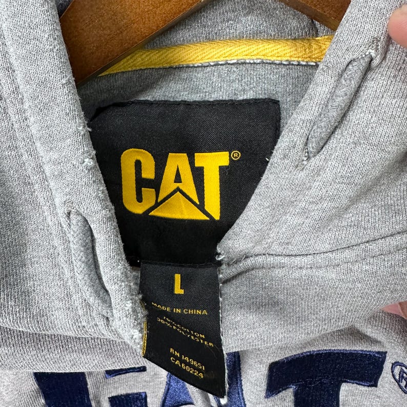 May include: Close-up of a gray hooded sweatshirt with the CAT logo in yellow and black. The size tag indicates a size L. The sweatshirt is made in China and is a blend of cotton and polyester.