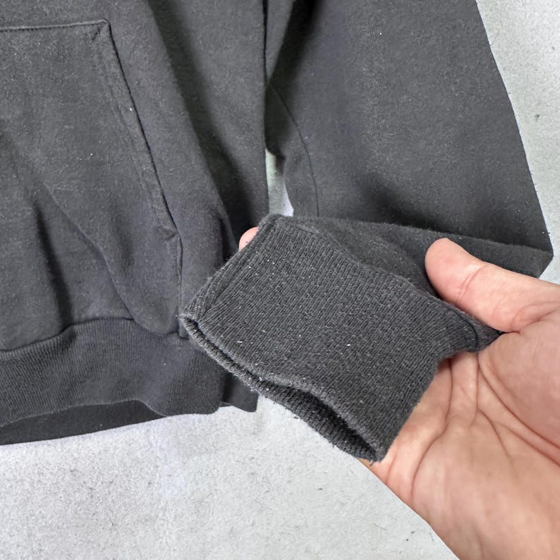 May include: Close-up of a black hooded sweatshirt. The image focuses on the sleeve cuff, showcasing the ribbed knit texture. The fabric appears slightly worn, with a visible pocket on the front. The overall style suggests casual wear.